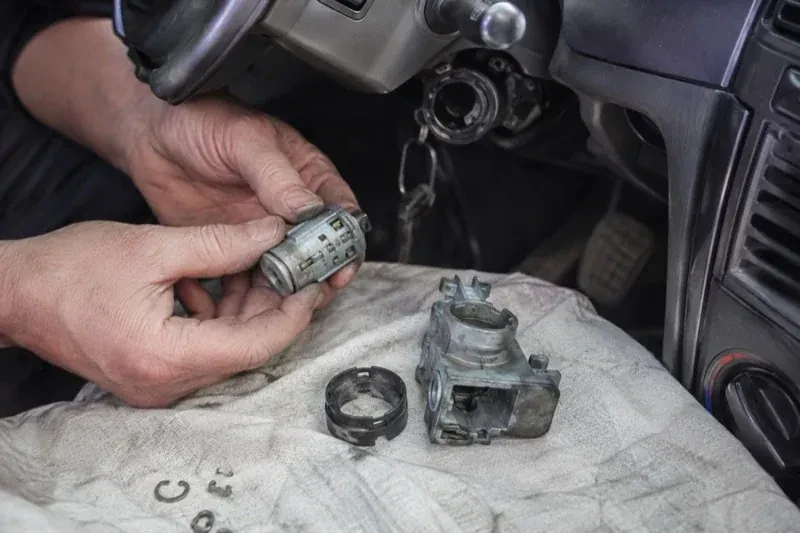 Hands holding a removed ignition cylinder beside a car steering column, with the housing and small parts on a cloth. — Ignition Lock Repair, Brantford
