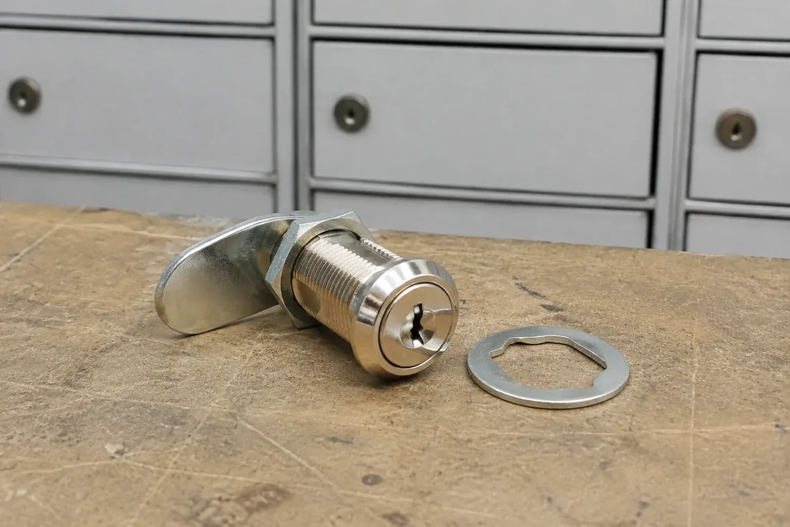 Detached threaded cam lock and mounting washer on a workbench in front of closed mail compartments. — Mailbox Lock Replacement, Brantford